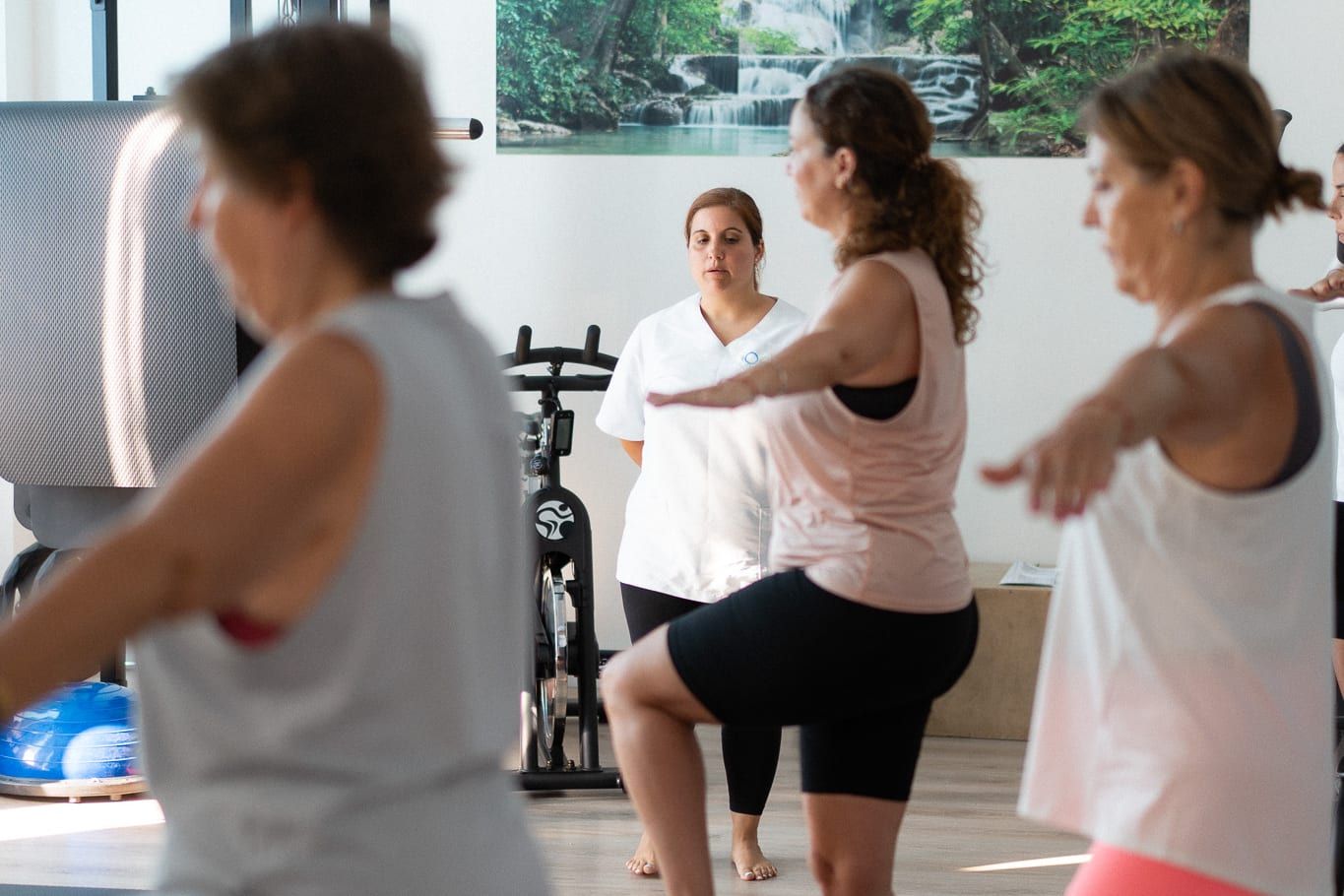 Alumnas de pilates hacen equilibrio sobre una pierna mientras la profesional las observa al fondo.